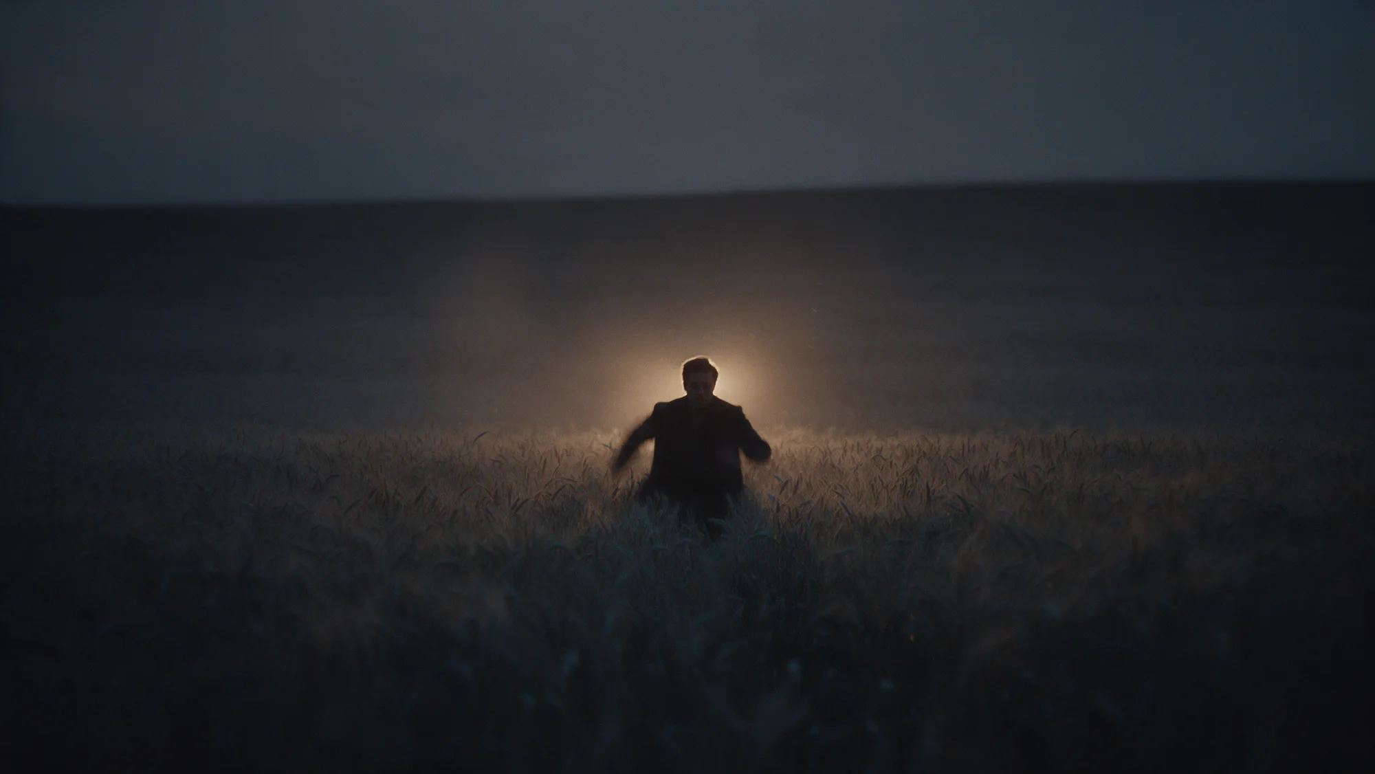 A man runs through a wheat field toward camera at dusk.