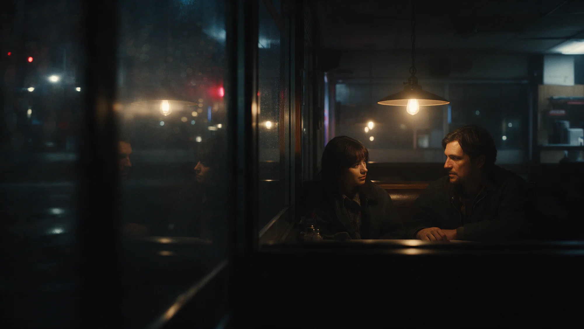 Two people across a late-night diner booth, seen through rain on the window.